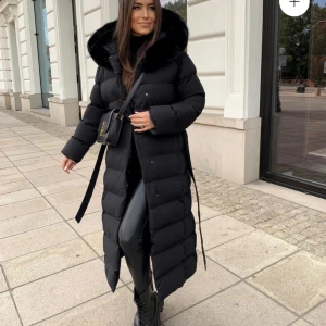 Svart dunjacka med pälsdetaljer - Snygg och varm svart dunjacka med pälsdetaljer på huvan. Jackan har en lång design med bälte och knappar som går att stänga. Den är nästintill oanvänd och har inga synliga defekter.