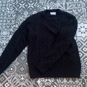 Svart stickad tröja från New Garments - Säljer en snygg svart stickad tröja från New Garments. Tröjan har ett klassiskt flätmönster och långa ärmar med ribbade muddar. Perfekt för kyligare dagar. Storlek L passar även M. Hör av dig vid intresse!