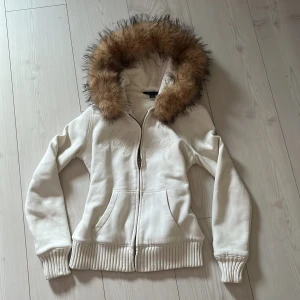 Vit zip up hoodie med fuskpäls från Tommy Hilfiger - Säljer en snygg vit zipup hoodie från Tommy Hilfiger med fuskpäls på huvan. Jackan har dragkedja framtill och broderad text på bröstet. Perfekt för kyliga dagar!! Sitter som en S