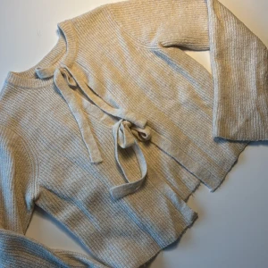 Beige stickad tröja från Object - Säljer en beige stickad tröja från Object med knytband framtill. Tröjan har långa ärmar och en ribbad struktur och två knytningar på framsidan så den både kan has som en tröja med två rosetter och en öppen kofta. Väldigt mysigt men också lite extra festlig🤗🫶