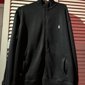 Ralph Lauren tröja - Säljer en stilren svart tröja från Ralph Lauren med dragkedja och det klassiska logotypen på bröstet. Tröjan har långa ärmar och ribbade muddar vid ärmslut och nederkant. Perfekt för en avslappnad stil. Den har dock någon problem med zippern men kan fortfarande zippas upp.