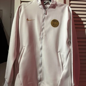 Nike psg tröja - Snygg vit Psg tröja från Nike med ett Paris-emblem på bröstet och guldfärgad Nike-logga. Jackan har dragkedja framtill och långa ärmar. Perfekt för en sportig look!