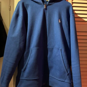 Blå hoodie från Ralph Lauren - Säljer en snygg blå hoodie från Ralph Lauren med dragkedja och huva. Tröjan har långa ärmar och en liten logga på bröstet. Perfekt för en avslappnad stil. Hör av dig vid intresse!