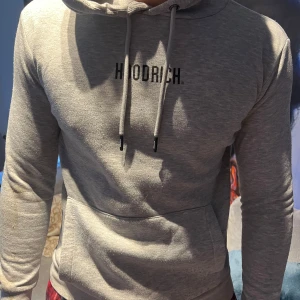 Grå hoodie från Hoodrich - Säljer en stilren grå hoodie från Hoodrich med justerbar huva och dragkedja på sidan. Perfekt för en avslappnad look. Den har långa ärmar och en bekväm passform.