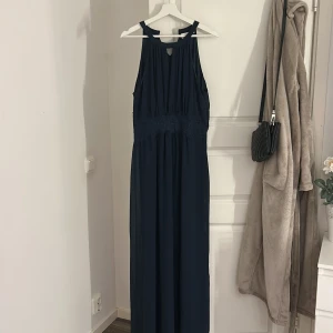 Mörkblå långklänning från Vila - Elegant mörkblå långklänning från Vila med vackra spetsdetaljer i midjan. Klänningen har en stilren design med en hög hals och är ärmlös, vilket ger en sofistikerad look. Perfekt för speciella tillfällen.