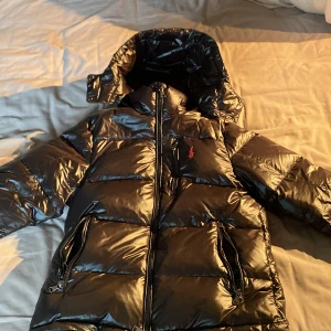 Svart dunjacka från Ralph Lauren - Säljer en snygg svart dunjacka från Ralph Lauren med röd broderad logga på bröstet. Jackan har en glansig finish och är utrustad med dragkedja och huva för extra skydd mot kyla. Perfekt för kalla dagar!