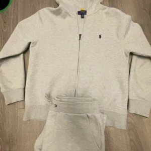 Grå tracksuit Ralph Lauren - Säljer en stilren grå tracksuit från Ralph Lauren med dragkedja och det klassiska logotypbroderiet på bröstet. Perfekt för en avslappnad look. Hoodien har långa ärmar och ribbade muddar.