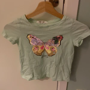 Mintgrön t-shirt med fjärilspaljetter från H&M - Säljer en söt mintgrön t-shirt från H&M med en stor fjäril i glittriga paljetter på framsidan. T-shirten har korta ärmar och är perfekt för en färgglad look. Passar barn i storlek 122/128. Men är stor i storleken så kan likväl passa de som har 140-146 
