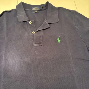 Mörkblå pikétröja från Ralph Lauren - Säljer en klassisk mörkblå pikétröja från Ralph Lauren med den ikoniska gröna loggan broderad på bröstet. Tröjan har en krage och knappar framtill. Perfekt för en stilren look.