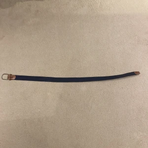 Blått flätat bälte med bruna detaljer - Snyggt blått flätat bälte med bruna skinnliknande detaljer och ett stilrent silverfärgat spänne. Perfekt för att ge en casual look lite extra stil. Passar bra till jeans eller chinos.