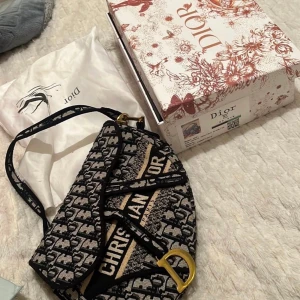 Svart och beige axelväska från Dior - Snygg axelväska från Dior i svart och beige med märkets logotypmönster. Väskan har guldfärgade metalldetaljer och en justerbar axelrem. Perfekt för att ge en lyxig touch till din outfit.