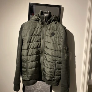 Grön jacka med huva - Snygg grön jacka med huva och dragkedja framtill. Jackan har quiltade detaljer och ribbade muddar. Ärmarna har en praktisk ficka med dragkedja. Perfekt för kyligare dagar. Man kan också välja om man vill ha luva eller inte 