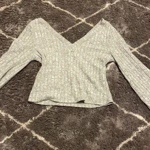 Grå ribbad omlottopp - Säljer en stilren grå ribbad omlottopp med v-ringning. Toppen har långa ärmar och en mjuk, bekväm passform. Perfekt för en avslappnad men chic look.