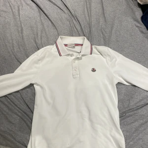 Vit pikétröja från Moncler - Säljer en stilren vit pikétröja från Moncler med lång ärm och klassisk krage. Tröjan har en broderad Moncler-logga på bröstet och knappar framtill. Perfekt för en avslappnad men ändå elegant look. säljer den för att den nu är för liten.