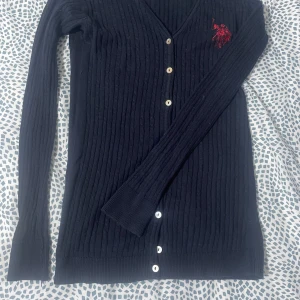 Svart ribbad kofta från U.S. Polo Assn. - Snygg svart ribbad kofta från U.S. Polo Assn. med knappar framtill och ett rött broderat emblem på bröstet. Perfekt för en stilren look. Långärmad design som passar bra till kyligare dagar.