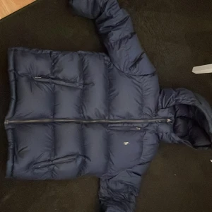 Mörkblå dunjacka från Ralph Lauren - Säljer en snygg mörkblå dunjacka från Ralph Lauren. Jackan har en praktisk dragkedja och en mysig huva. Perfekt för kyliga dagar med sin pufferdesign som håller dig varm och bekväm. Den har långa ärmar och två fickor framtill för extra funktionalitet. Bra skick förutom loggan.