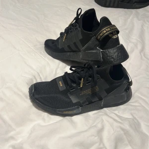 Svarta Adidas NMD sneakers med gulddetaljer - Snygga svarta Adidas NMD sneakers med gulddetaljer. Skorna har en modern design med text på hälen och snörning framtill. Perfekta för en stilren look.
