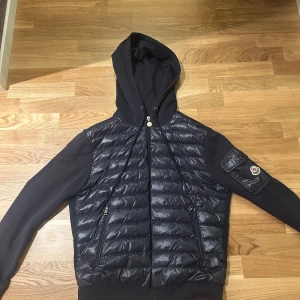 Moncler cardigan bra skick  - Säljer en snygg svart pufferjacka från Moncler med huva och dragkedja. Jackan har långa ärmar och två dragkedjefickor framtill. Perfekt för kyligare dagar.