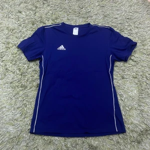 Blå t-shirt från Adidas - Säljer en blå t-shirt från Adidas med korta ärmar och vita detaljer längs sidorna. Perfekt för träning eller vardagsbruk. Adidas-loggan är tryckt på bröstet. Tillverkad i ett lätt och bekvämt material.