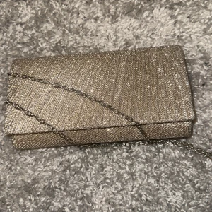 Glittrig kuvertväska med kedja - Snygg glittrig kuvertväska i silver med en elegant kedja. Perfekt för att ge din outfit en extra touch av glamour. Väskan har en stilren design och är lätt att bära med sig. Perfekt till balen. Använt endast en gång. 