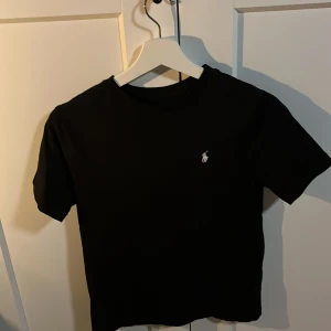 Svart t-shirt från Polo Ralph Lauren - Säljer en klassisk svart t-shirt från Polo Ralph Lauren med det ikoniska broderade logotypen på bröstet. Perfekt för en stilren look. T-shirten är kortärmad och gjord i mjukt material.