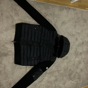  Moncler Cardigan - Säljer en snygg svart Cardigan från Moncler. Jackan har en quiltad design med dragkedja och huva. Perfekt för höst och vinter, håller dig varm och stilren. Den har långa ärmar och praktiska fickor med knappar. Passar både till vardags och lite finare tillfällen.