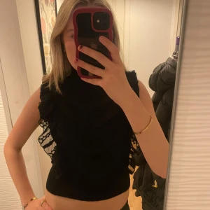 Svart topp med spetsdetaljer - Säljer en snygg svart topp med spetsdetaljer och volanger. Den är ärmlös och har en hög hals, vilket ger en elegant look. Perfekt för festliga tillfällen eller en utekväll. Toppen är croppad och sitter tight, vilket framhäver figuren.