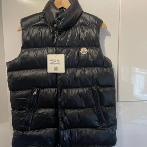 dunväst Moncler - Säljer en snygg svart dunväst från Moncler i oanvänt skick. Västen har en hög krage och dragkedja framtill, perfekt för kyliga dagar. Rfid koden fungerar som den ska. Säljes pga behov av pengar! Storlek 2. Motsvarar Medium 