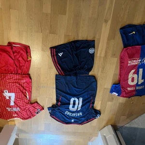 Fotbollströjor från olika lag - Säljer flera fotbollströjor i olika färger och stilar. En röd och gul randig tröja, en blå och röd tröja, en svart tröja med röda detaljer, en rosa tröja och en svart och röd randig tröja. Perfekt för fotbollsfans som vill ha en bit av sina favoritlag! Alla tröjor är i bra skick och redo för match eller samling.