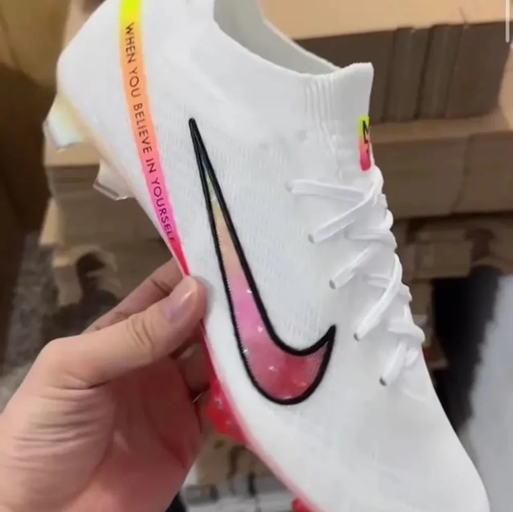 Snygga vita Nike Mercurial fotbollsskor med regnbågsfärgad sula och texten 'When you believe in yourself' på sidan. Skorna har en modern design med snörning och en stilren svart Nike-logga. Perfekta för att sticka ut på planen!. Skor.