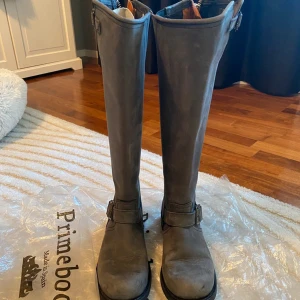 Grå Primeboots i skinn - Säljer ett par snygga grå Primeboots i mockaskinn att 37. De har en hög modell med dragkedja baktill och dekorativa spännen. Perfekta för höst och vinter, och de är i mycket bra skick. Passar både till vardags och lite finare tillfällen. Dyra i inköp men säljs billigt då de bara står i garderoben