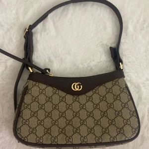 Brun axelväska med monogram - Säljer en snygg axelväska i mycket bra skick. Väskan har ett klassiskt monogrammönster och guldfärgade detaljer. Den är perfekt för både vardag och fest. Väskan har en dragkedja upptill och justerbar axelrem. En tidlös accessoar för alla tillfällen!