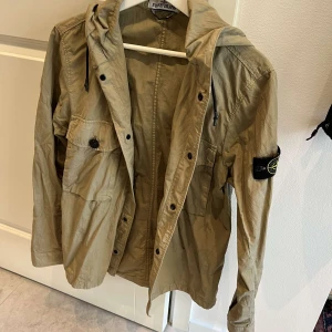 Beige jacka från Stone Island - Säljer en snygg beige jacka från Stone Island i mycket bra skick. Jackan har en huva och långa ärmar. Perfekt för höst och vår, med knappar och dragkedja framtill. Passar både till vardags och lite mer uppklädda tillfällen. Den är knappt använd och är i storlek S. 