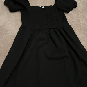 Svart klänning med puffärmar - Säljer en elegant svart klänning med puffiga kortärmar och smockdetaljer på överdelen. Perfekt för både vardag och festliga tillfällen. Klänningen har en bekväm passform och är gjord i ett mjukt material som faller fint.