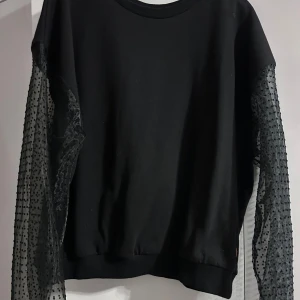 Svart tröja med prickiga meshärmar - Snygg svart tröja med unika prickiga meshärmar som ger en cool twist. Perfekt för både vardag och fest! Tröjan har en normal passform och är i mycket bra skick. Testad men aldrig använd 