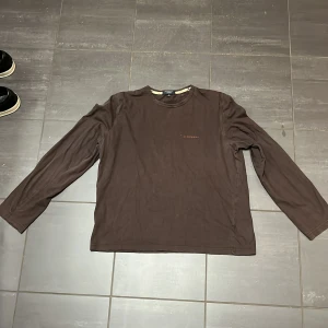 Burberry - Säljer en stilren brun långärmad tröja från Burberry i bomull. Perfekt för höst och vinter med klassisk design och bekväm passform. Tröjan har en diskret logga på bröstet och den ikoniska Burberry-rutiga insidan av kragen. Passar både till vardags och lite finare tillfällen.