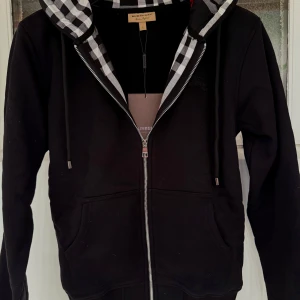 Burberry Hoodie - Säljer en snygg svart hoodie från Burberry med klassiskt rutigt mönster i luvan. Den har dragkedja framtill och praktiska fickor. Perfekt för höst och vår, både stilren och bekväm. Passar till alla tillfällen! Obs:skriv innan köp!