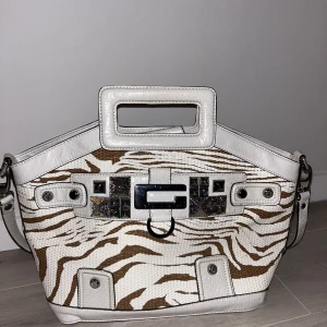 Vit och brun axelväska med zebramönster - Säljer denna axelväska från märket Guess i vitt och brunt med coolt zebramönster. Väskan har en praktisk dragkedja och ett stilrent handtag. Säljer pga att den inte kommer till användning