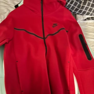 Nike tech  - Säljer en snygg röd hoodie från Nike i mycket bra skick. Den har en dragkedja framtill och långa ärmar. Perfekt för höst och vår, och funkar lika bra till träning som till vardags. Den har en cool design med svarta detaljer och en praktisk ficka på ärmen.