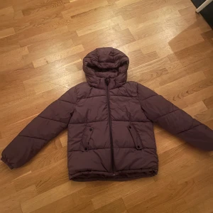 Lila pufferjacka från H&M - Säljer en snygg lila pufferjacka från H&M i storlek EUR 158. Jackan har en mysig huva och praktiska fickor med knappar. Perfekt för kyliga höst- och vinterdagar. Den är i bra skick och redo för nya äventyr!