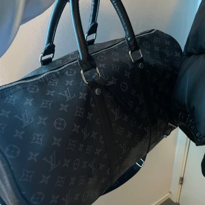 Svart resväska med monogrammönster  - Snygg svart resväska med klassiskt monogrammönster. Perfekt för resor med stil! Väskan har en dragkedja och justerbar axelrem för bekväm bärning. Den är rymlig och passar perfekt för en weekendresa eller som handbagage.