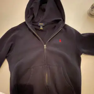 Säljer en snygg blå hoodie från Ralph Lauren med dragkedja och det klassiska röda logotypen på bröstet. Perfekt för höst och vinter, den är både stilren och bekväm. Passar till alla tillfällen, oavsett om du ska chilla hemma eller ut på stan. 9/10 skick