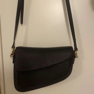 Svart axelväska i skinn - Snygg svart axelväska i skinn med en stilren design. Väskan har en justerbar axelrem och en praktisk dragkedja inuti för extra säkerhet. Perfekt för både vardag och fest!