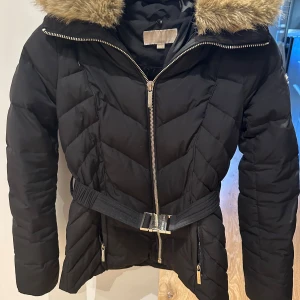 Svart dunjacka från Michael Kors - Säljer en snygg svart dunjacka från Michael Kors i storlek XS. Jackan har en mysig fuskpäls på kragen och ett bälte i midjan för en fin passform. Den är perfekt för kalla vinterdagar och har en dragkedja framtill. Jackan är i mycket bra skick och redo för nya äventyr!