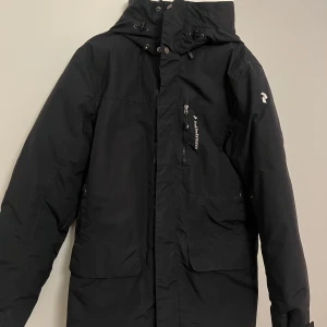 Svart vinterjacka från Peak Performance - Säljer en svart vinterjacka från Peak Performance i mycket bra skick. Jackan har en praktisk dragkedja och knappar framtill, samt en justerbar huva. Perfekt för kalla vinterdagar med sin varma och tåliga design. Storleken är herr S