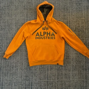 Orange hoodie från Alpha Industries - Säljer en snygg orange hoodie från Alpha Industries i storlek S. Den har en stor logga på framsidan. Perfekt för höst och vår med sin mjuka och bekväma passform. Huvan har justerbara snören för extra komfort. Passar både till vardags och chillkvällar. 🧡