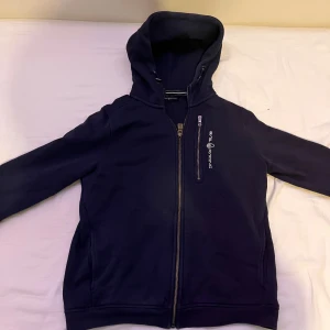 Mörkblå hoodie från sail racing - Säljer en snygg mörkblå hoodie från sail racing. Den har en dragkedja framtill och en bekväm huva. Perfekt för kyliga dagar och passar till det mesta. Den är i bra skick och redo för en ny ägare!