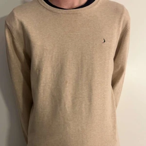 Beige tröja med liten logga - Säljer en stilren beige tröja med en liten diskret logga på bröstet. Tröjan har långa ärmar och är gjord i ett mjukt material som känns skönt mot huden. Perfekt för både vardag och lite finare tillfällen. Passar bra till jeans eller chinos!