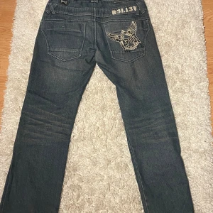 Jeans från Tapout - Säljer ett par coola jeans från Tapout med tryck på både bakfickan och benet. De är mörkblå och har en avslappnad passform. Perfekta för en casual look eller för att sticka ut lite extra. Passar bra till vardags eller för en chill kväll med vänner.