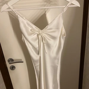 Vit satin klänning från BikBok - Säljer en elegant vit satin klänning från BikBok i storlek XS. Klänningen har smala axelband och en v-ringad design med knytning framtill. Perfekt för sommarens fester eller en speciell kväll. Den är i nytt skick och ger en lyxig känsla.